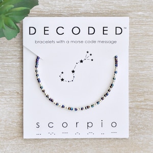 May include: A beaded bracelet with a silver and purple color scheme. The bracelet features a constellation of stars representing the zodiac sign Scorpio. The card reads "DECODED" and "bracelets with a morse code message" and "Scorpio" with the morse code for the word.