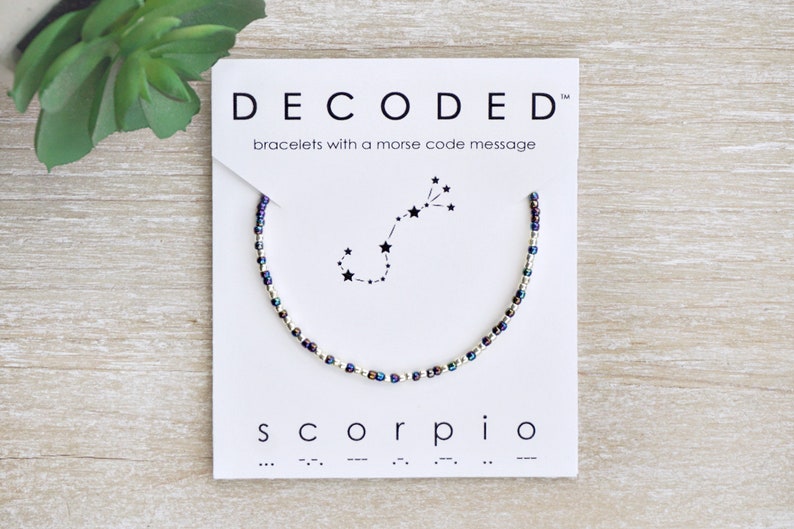 May include: A beaded bracelet with a silver and purple color scheme. The bracelet features a constellation of stars representing the zodiac sign Scorpio. The card reads "DECODED" and "bracelets with a morse code message" and "Scorpio" with the morse code for the word.