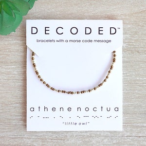 May include: A beaded bracelet with a Morse code message, displayed on a white card. The card reads "DECODED" and "athene noctua" with the phrase "little owl" below. The bracelet features gold and brown beads.