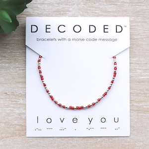 May include: A red and silver beaded bracelet on a white card. The card reads "DECODED" and "bracelets with a morse code message." Below the bracelet, the card says "love you" in text and Morse code.