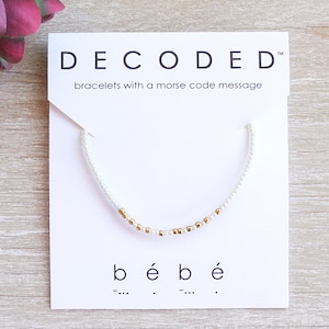 May include: White beaded bracelet with gold beads spelling out "bébé" in Morse code on a white card with the text "DECODED™ bracelets with a morse code message"