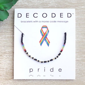 May include: A black beaded bracelet with a rainbow colored stripe and gold beads. The bracelet is on a white card with the text "DECODED" and "bracelets with a morse code message". The card also has a rainbow ribbon and the word "pride" in morse code.