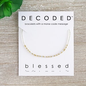 May include: A white card with the text "DECODED" and "bracelets with a morse code message" printed on it. A silver bracelet with gold beads is displayed on the card. The word "blessed" is printed below the bracelet, along with the morse code for the word.