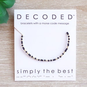 May include: A black and gold beaded bracelet with the text "DECODED" and "bracelets with a morse code message" on a white card. The bracelet has the text "simply the best" in morse code.