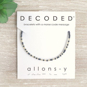 May include: A black and gold beaded bracelet with the word "allons-y" and its Morse code translation on a white card. The card also reads "DECODED" and "bracelets with a morse code message".