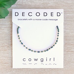May include: A green and gold beaded bracelet with the word "cowgirl" and its morse code translation on a white card. The card also reads "DECODED" and "bracelets with a morse code message".