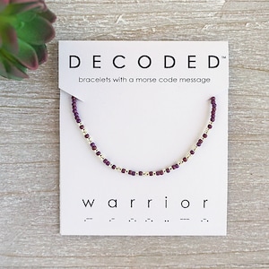 May include: A beaded bracelet with a Morse code message, displayed on a white card. The bracelet features small, dark purple beads and gold-colored spacers. The card reads "DECODED" and "warrior".