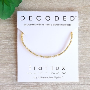 May include: A gold and silver beaded bracelet with the message "fiat lux" in morse code. The bracelet is on a white card with the text "DECODED™ bracelets with a morse code message".