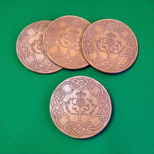 May include: Four circular, light brown coasters featuring a Celtic knot design and a thistle motif. The coasters are displayed on a green surface, highlighting the detailed carved patterns. Each coaster is approximately 10 cm in diameter.