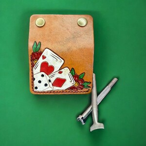 May include: A tan leather card holder with a playing card and dice design, featuring red hearts and diamonds. The holder has two brass snaps and is accompanied by two silver tools. The design includes red flowers and green leaves.