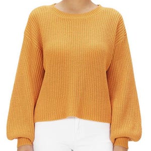 May include: Mustard yellow knit sweater with a round neckline and long, voluminous sleeves. The sweater is paired with white pants. The knitwear has a ribbed texture and a relaxed fit, suitable for casual wear.