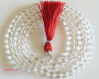 Puede incluir: Un collar mala de cristal de cuarzo transparente con una borla roja brillante. Las cuentas redondas están ensartadas, formando un patrón circular. La borla está atada con alambre plateado. El collar se muestra sobre una superficie blanca.