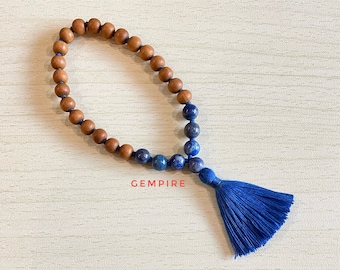 May include: A wooden and lapis lazuli beaded bracelet with a blue tassel. The bracelet has a brown wooden bead section and a blue lapis lazuli bead section. The tassel is made of blue fabric.