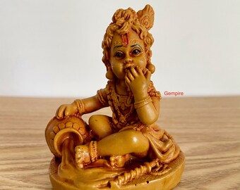 May include: A small, brown figurine of a young boy sitting with a pot of water pouring out. The boy is wearing a beaded necklace and a crown. The figurine is made of resin and is about 4 inches tall.