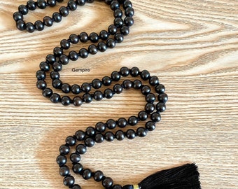 Puede incluir: Un collar de cuentas mala de madera negra con un borla negra. El collar está hecho de cuentas redondas y tiene un acento dorado al final de la borla.