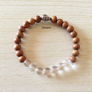 Könnte beinhalten: Ein braunes Holzperlenarmband mit einem silbernen Buddha-Anhänger und klaren Quarzperlen. Das Armband liegt auf einer hellbraunen Holzoberfläche. Gempire.
