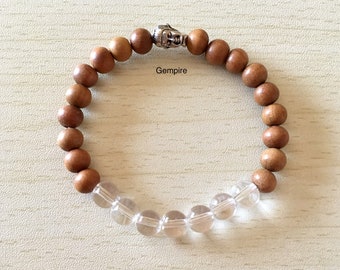 May include: A brown wooden bead bracelet with a silver Buddha charm and clear quartz beads. The bracelet is on a light wood surface. Gempire.