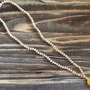 Pu&ograve; includere: Una lunga collana di perline bianco sporco con una nappa gialla. Le perline sono infilate su un cordoncino di colore chiaro. La collana &egrave; esposta su una superficie di legno scuro e strutturato. La parola "Gempire" &egrave; visibile.