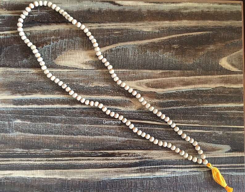 Pu&ograve; includere: Una lunga collana di perline bianco sporco con una nappa gialla. Le perline sono infilate su un cordoncino di colore chiaro. La collana &egrave; esposta su una superficie di legno scuro e strutturato. La parola "Gempire" &egrave; visibile.
