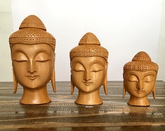 May include: Three wooden Buddha head sculptures of varying sizes. The sculptures are carved in a light brown color and have closed eyes. The sculptures are arranged in a row on a wooden surface.