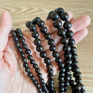 May include: Four strands of black wooden beads, each with a different size: 6 mm, 8 mm, 10 mm, and Gempire. The beads are strung on a cord and are displayed on a hand.