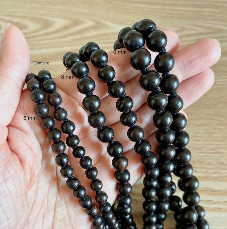 May include: Four strands of black wooden beads, each with a different size: 6 mm, 8 mm, 10 mm, and Gempire. The beads are strung on a cord and are displayed on a hand.