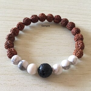 May include: A brown and white beaded bracelet with a black lava rock bead in the center. The bracelet is made with rudraksha beads and white howlite beads.