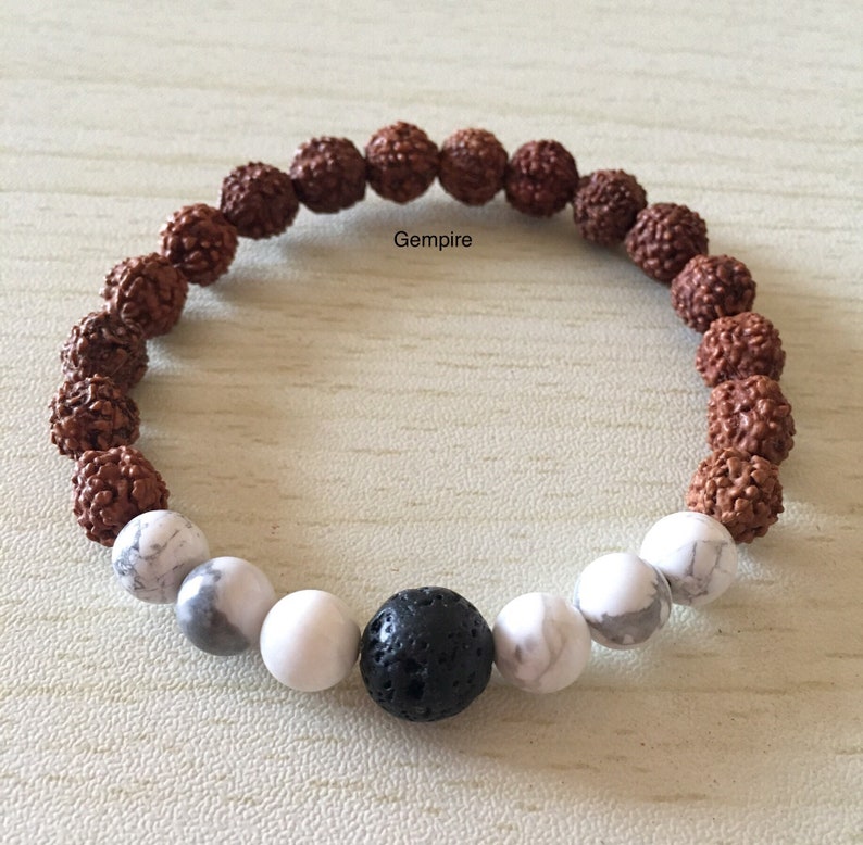 May include: A brown and white beaded bracelet with a black lava rock bead in the center. The bracelet is made with rudraksha beads and white howlite beads.