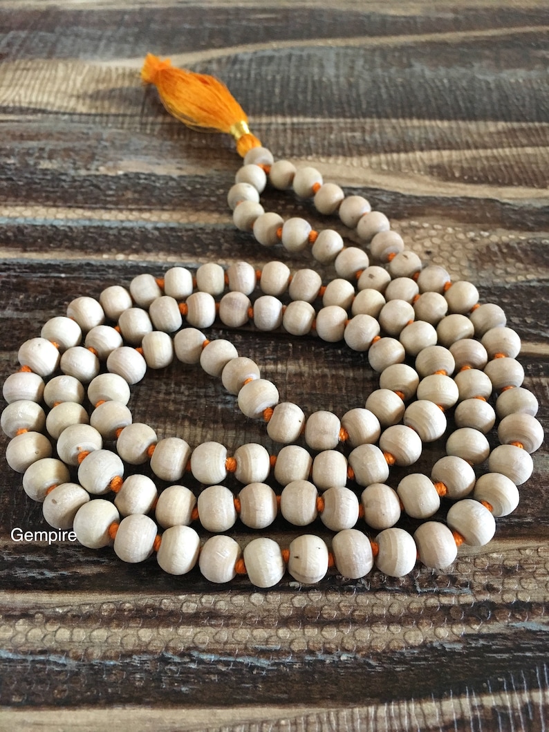 Pu&ograve; includere: Una collana di perline mala in legno con perline di colore chiaro infilate su filo arancione. La collana ha una nappa arancione brillante all'estremit&agrave;. Le perline sono lisce e rotonde. La collana &egrave; esposta su una superficie di legno.