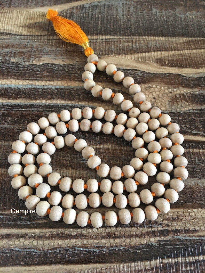 Pu&ograve; includere: Una collana di perline mala in legno color chiaro con un nappa arancione. Le perline sono infilate su un filo arancione. La parola "Gempire" &egrave; stampata in basso a sinistra nell'immagine.