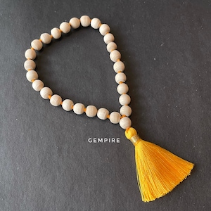 May include: A white wooden beaded necklace with a yellow tassel. The necklace is strung on a thin orange thread. The tassel is made of a soft, fluffy material.