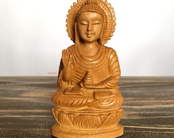 May include: A hand-carved wooden statue of Buddha in a seated meditation pose. The statue is light brown in color and has a serene expression. The statue is sitting on a lotus flower base.