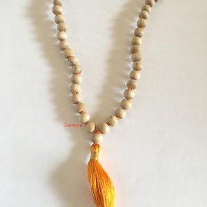 Pu&ograve; includere: Una collana di perline di legno color chiaro con una nappa arancione. Le perline sono rotonde e infilate su un filo arancione. La nappa &egrave; giallo brillante e ha una fascia color oro. La parola "Gempire" &egrave; stampata sull'immagine.
