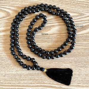 May include: A black wooden mala bead necklace with a black tassel. The beads are 10 mm in diameter. The necklace is on a wooden surface. Gempire
