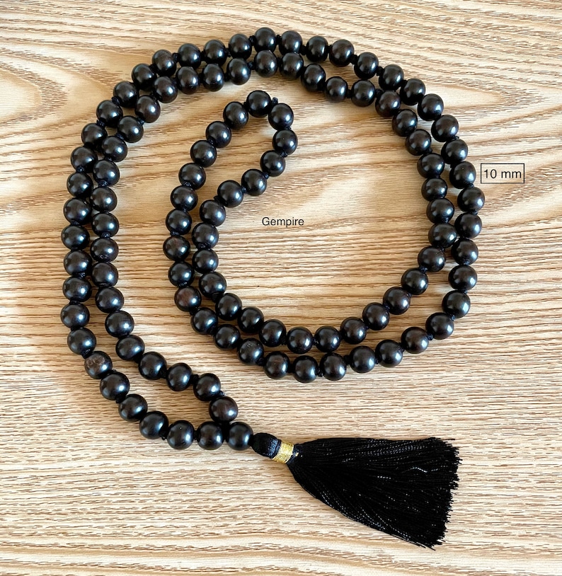 May include: A black wooden mala bead necklace with a black tassel. The beads are 10 mm in diameter. The necklace is on a wooden surface. Gempire