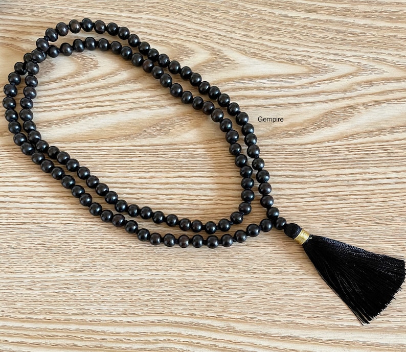 May include: A black wooden mala bead necklace with a black tassel. The necklace is made of 108 beads and is approximately 18 inches long.