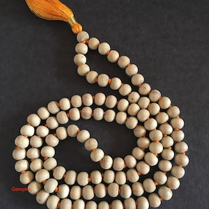 Pu&ograve; includere: Una collana di perline di legno color chiaro con un nappa e un filo arancione. Le perline sono di forma ovale e infilate con filo arancione. La nappa &egrave; fatta di fili arancioni e filo dorato.