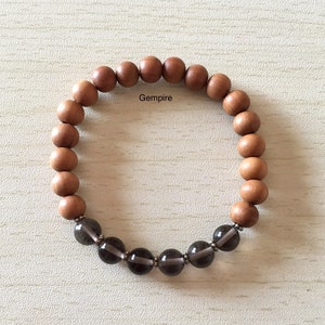 Puede incluir: Un brazalete de cuentas de madera marrón con una fila de cuentas de piedras preciosas gris oscuro. El brazalete tiene la palabra "Gempire" impresa.
