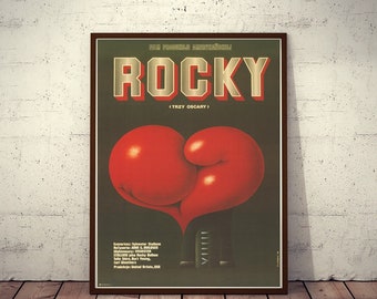Puede incluir: Un póster de película vintage para la película "Rocky" con un fondo verde y una gran forma de corazón de guante de boxeo rojo. El título "ROCKY" está en letras doradas con el texto "(TRZY OSCARY)" debajo. El póster también incluye el texto "Scenariusz: Sylvester Stallone" y "Relyseria: John G. Avildsen".