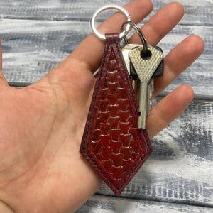 Op de afbeelding: Een rode leren sleutelhanger met een geometrisch patroon en een zilveren sleutelring. De sleutelhanger is ruitvormig en bevat meerdere sleutels. Het leer heeft een getextureerde uitstraling.