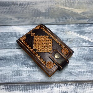 Puede incluir: Cartera de cuero marrón oscuro con intrincados diseños en relieve dorados. La cartera tiene forma rectangular con cierre de botón a presión y patrones florales decorativos. El fondo es una superficie de madera azul claro y gris.