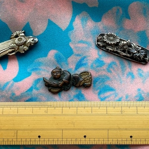May include: Three antique metal decorative objects. One is a miniature piano, another is a rectangular bar with floral designs, and the third is a small figure of a person reading. The objects are on a blue and pink patterned fabric.