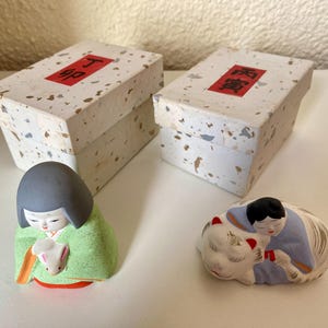 May include: Two decorative boxes with red text sit behind two Japanese figurines. One figurine is a girl in a green kimono holding a small white rabbit. The other figurine is a person in a blue kimono hugging a white cat.