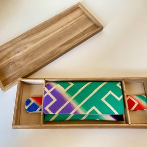 May include: A rectangular wooden box with a removable lid, containing a folded fabric item featuring a geometric pattern in shades of green, purple, and white, with additional fabric accents in red, blue, and white visible on the sides.