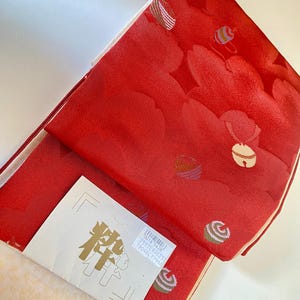 May include: A folded red textile with a repeating pattern of stylized bell shapes and other designs. A white tag with gold Japanese characters and a barcode is visible. The textile has a cream-colored lining.