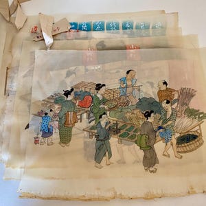 May include: A stack of vintage Japanese woodblock prints depicting a bustling market scene. The prints feature figures in traditional attire, surrounded by produce and market stalls. The prints have a faded, aged appearance.
