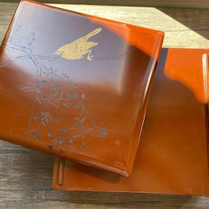 Puede incluir: Una caja rectangular de color marrón rojizo lacada con tapa. La tapa presenta un pájaro dorado posado en una rama con diseños grabados de flores y hojas. La caja está abierta, revelando el interior y la base.
