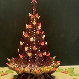 Könnte beinhalten: Ein Keramik-Weihnachtsbaum mit brauner Glasur, verziert mit roten und weißen Lichtern. Ein rosa Stern ziert die Spitze des Baumes. Der Sockel ist goldfarben und sternförmig. Der Baum wird von kleinen, warmen Lichtern beleuchtet.