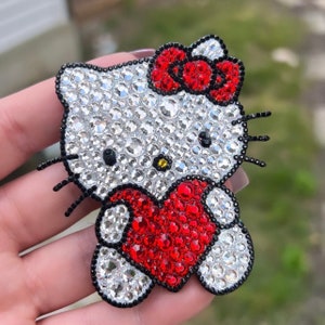 May include: A rhinestone-encrusted Hello Kitty charm with a red heart. The charm is made of clear and red rhinestones.
