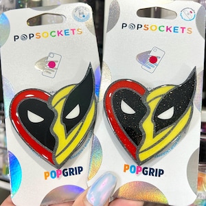 May include: Two PopSockets PopGrip phone grips featuring a red and yellow heart with a black and yellow Wolverine mask design. The text "POPGRIP" is printed on the packaging.